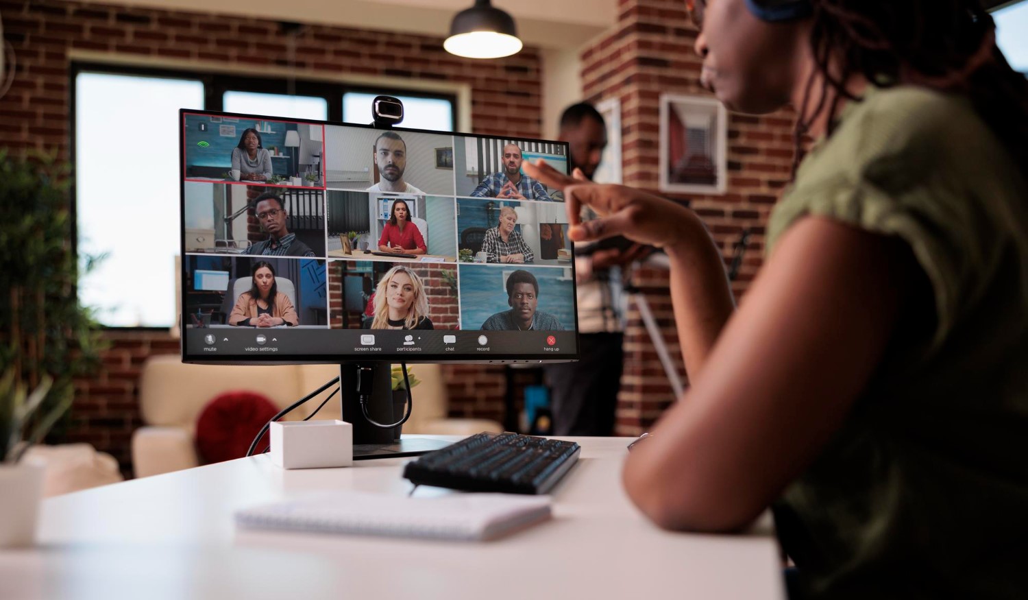 Person sitting at desk pointing at a computer monitor showing a video call with multiple participants in a modern office with brick walls and large windows